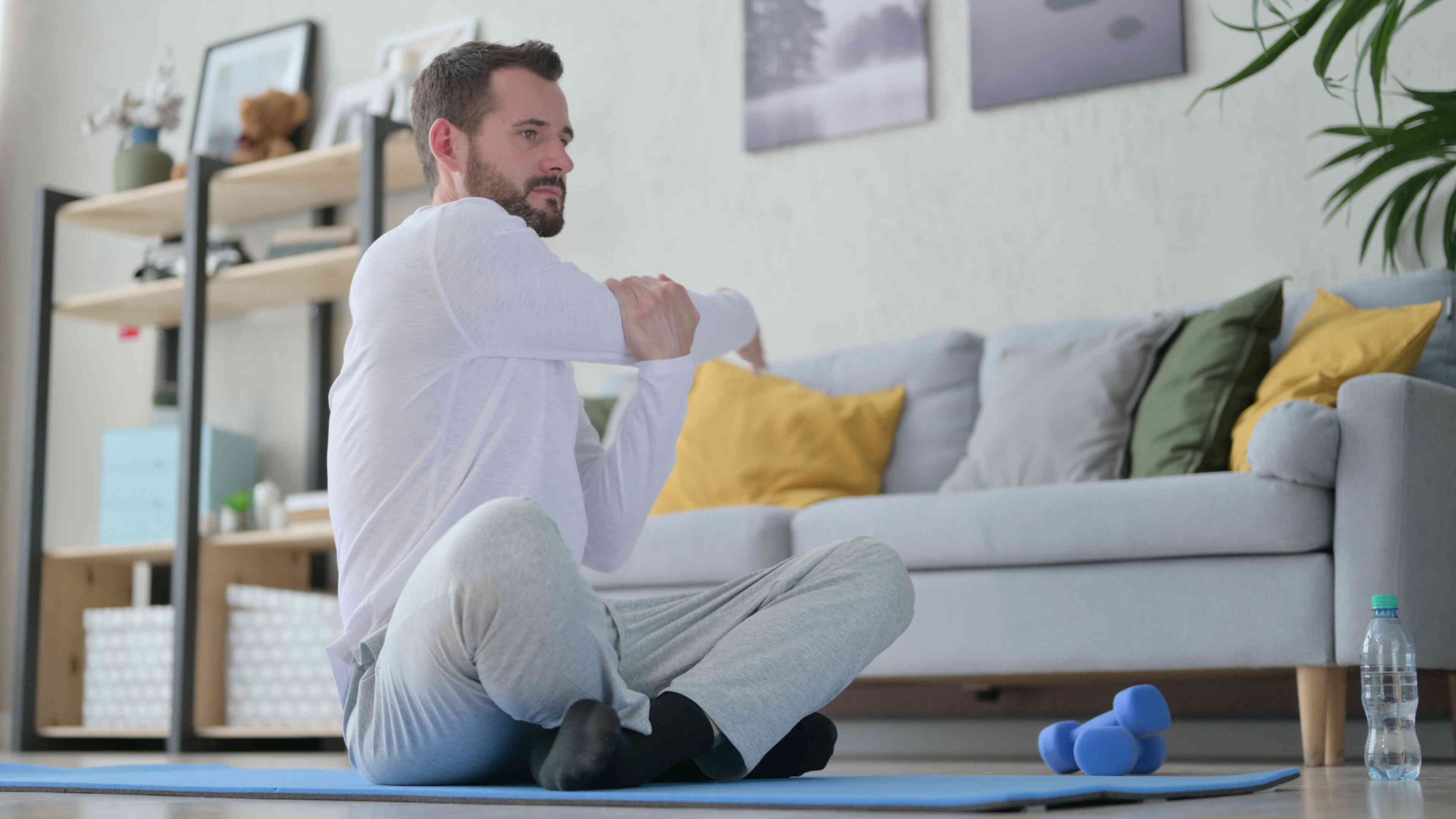 A man in a white shirt stretches his arms while sitting on a yoga mat on his living room floor.